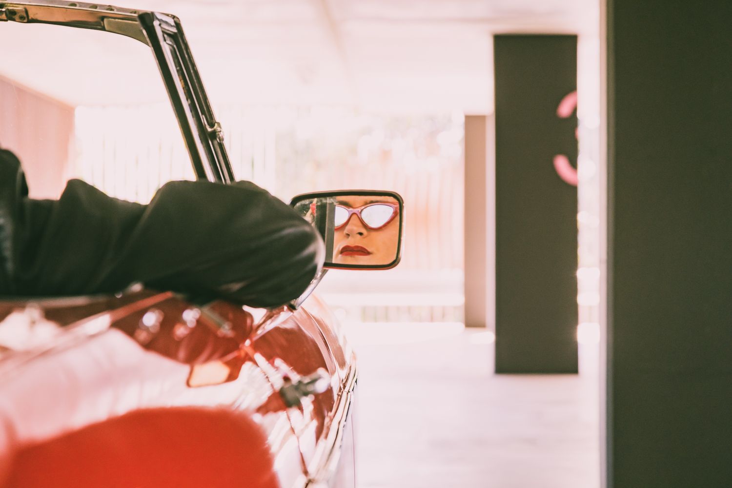 Person in a black jacket rests an arm on a red convertible, with sunglasses reflected in the side mirror.