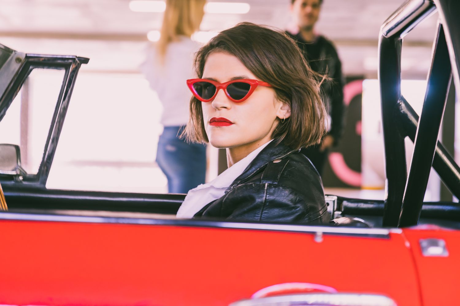 A woman in red cat-eye sunglasses, red lipstick and a black leather jacket sits in a vintage red convertible.