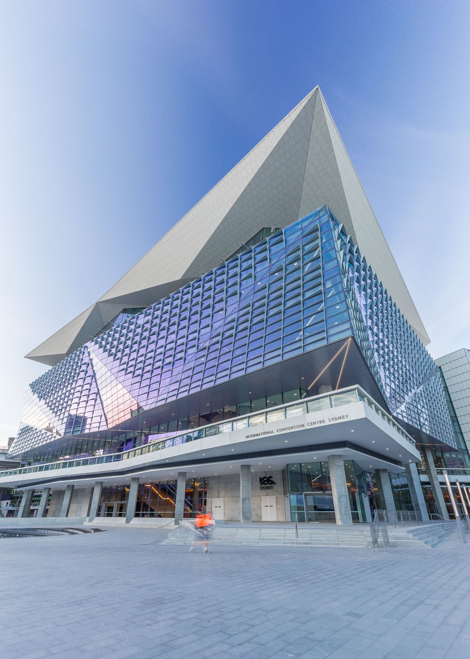 Striking angular convention centre with a pyramid-shaped white roof and shimmering blue glass facade rising above a paved plaza.