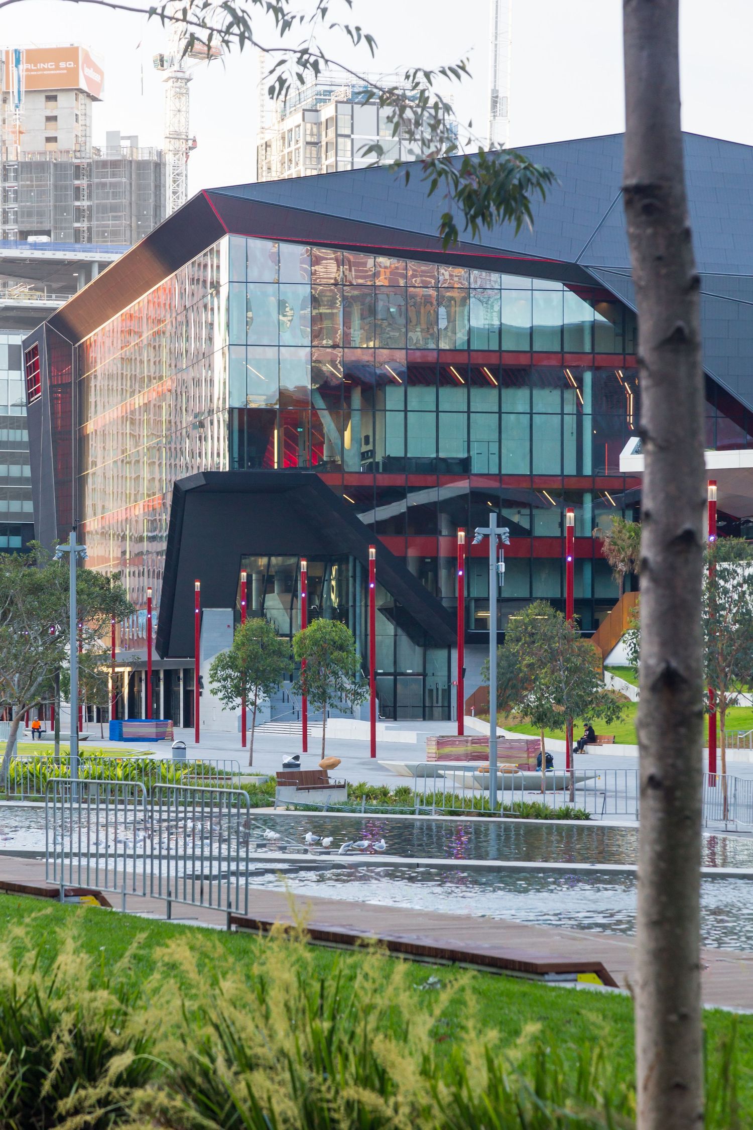 Glass and steel convention building with red columns and dark angular roof overlooking a water feature and trees in Sydney.
