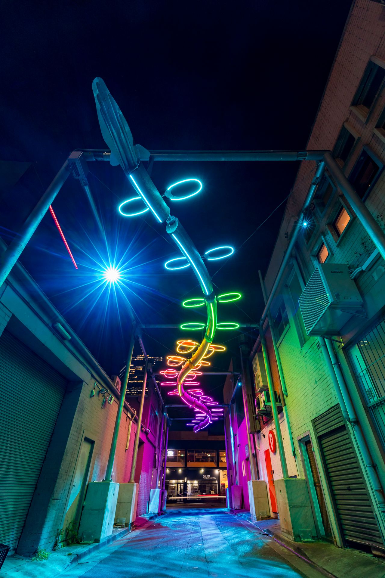 Colorful neon light installation in green, yellow and pink spiraling above a narrow urban laneway at night between brick buildings.