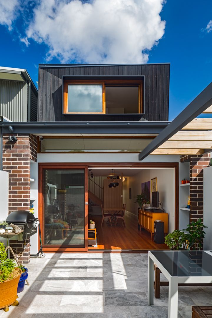 Modern two-storey home with a dark timber-clad upper level, brick base and open sliding doors leading to a sunny paved courtyard.