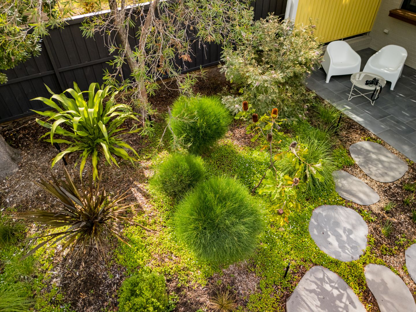 Garden with mounded green plants, spiky grasses and round stepping stones leading to white chairs on a patio.