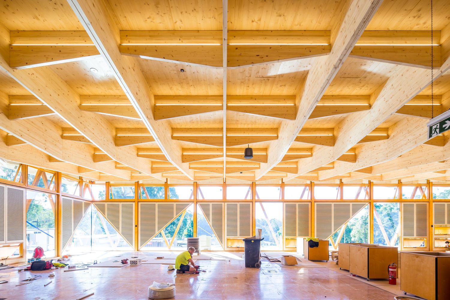 Interior of a timber building under construction with exposed wooden roof beams, large windows and workers on the floor.