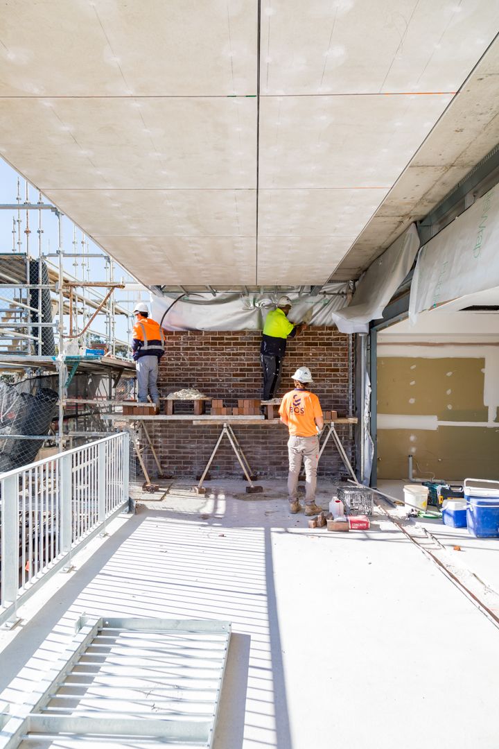 Construction workers in high-visibility clothing install brick and panel work on a building balcony with scaffolding nearby.