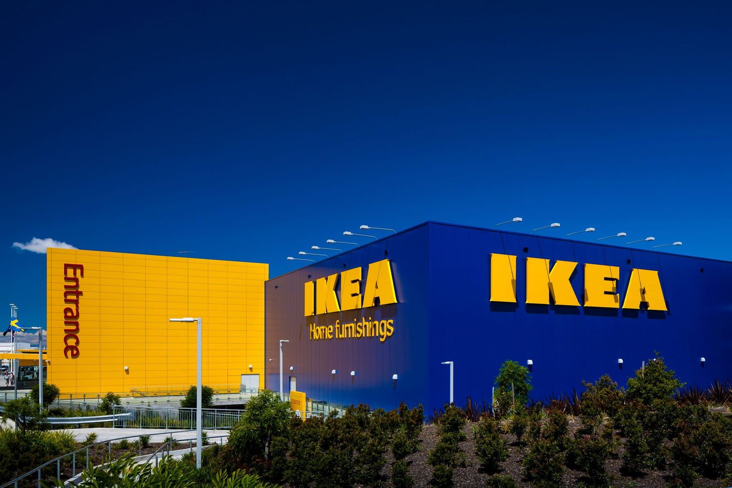 Blue and yellow IKEA store exterior with illuminated logo, entrance sign and landscaped trees under a clear blue sky.