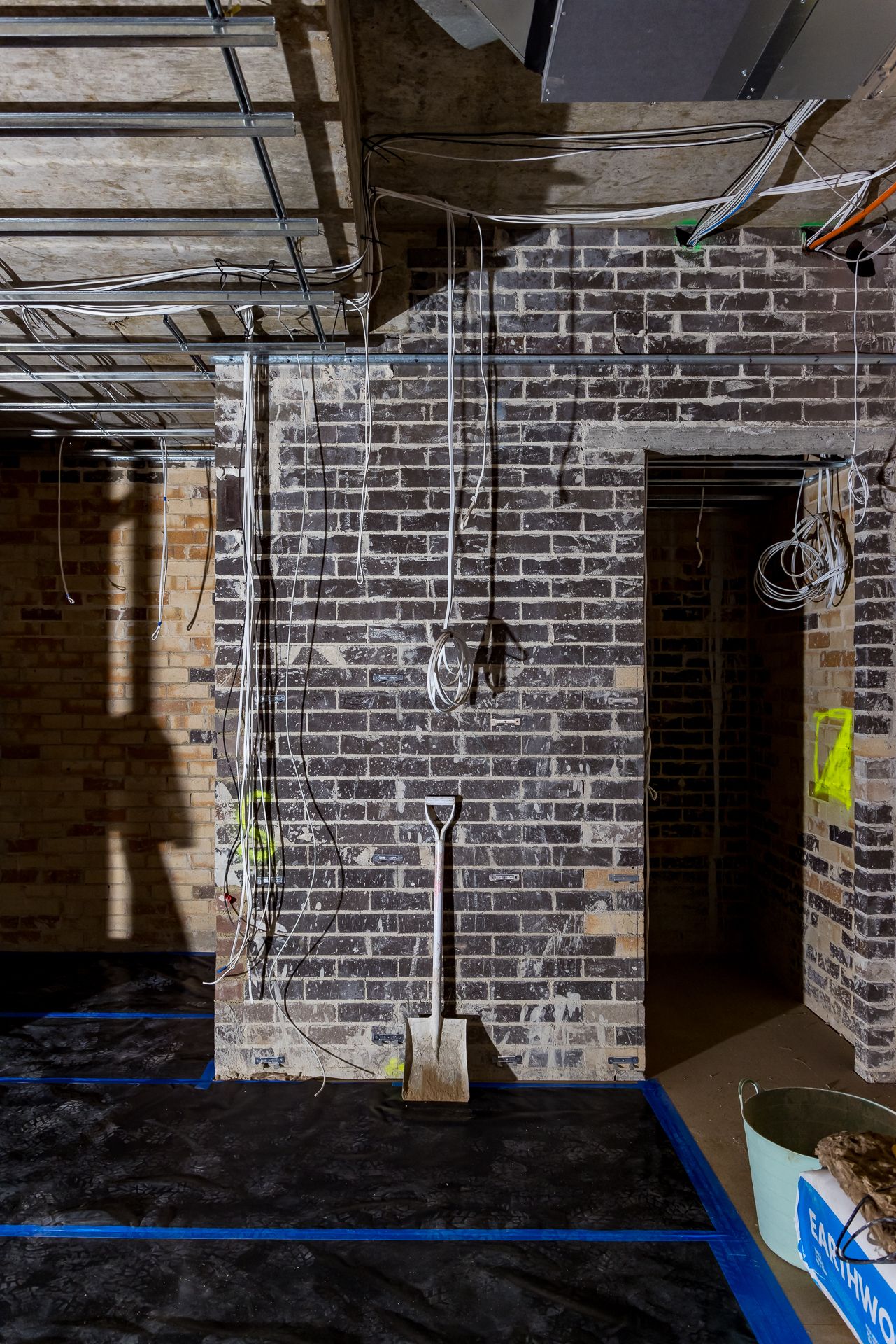 Construction interior with exposed dark brick walls, loose electrical wiring, a shovel leaning against the wall and blue floor tape.