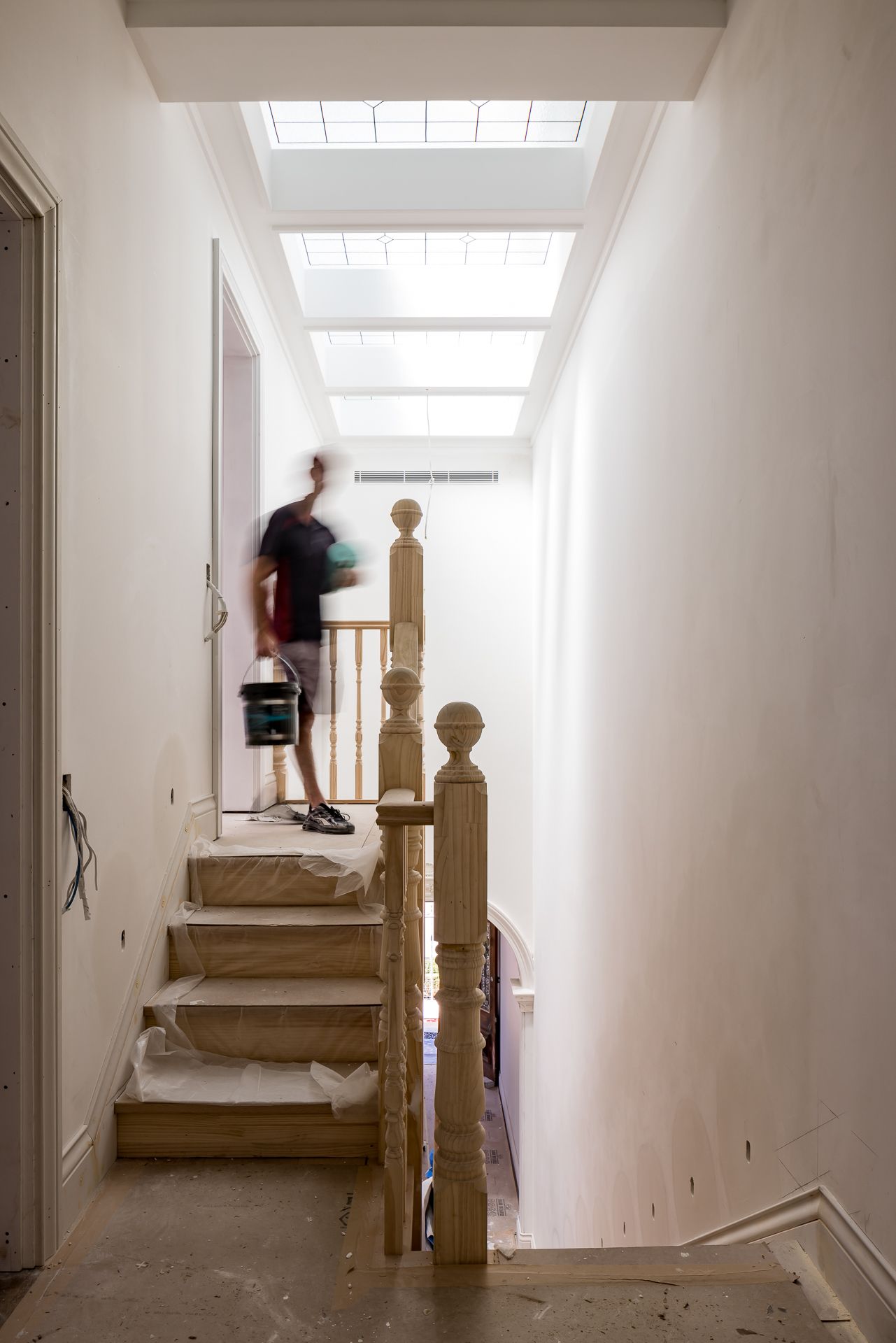 Blurred person carrying a paint bucket up a protected timber staircase in a renovated home with skylights above.