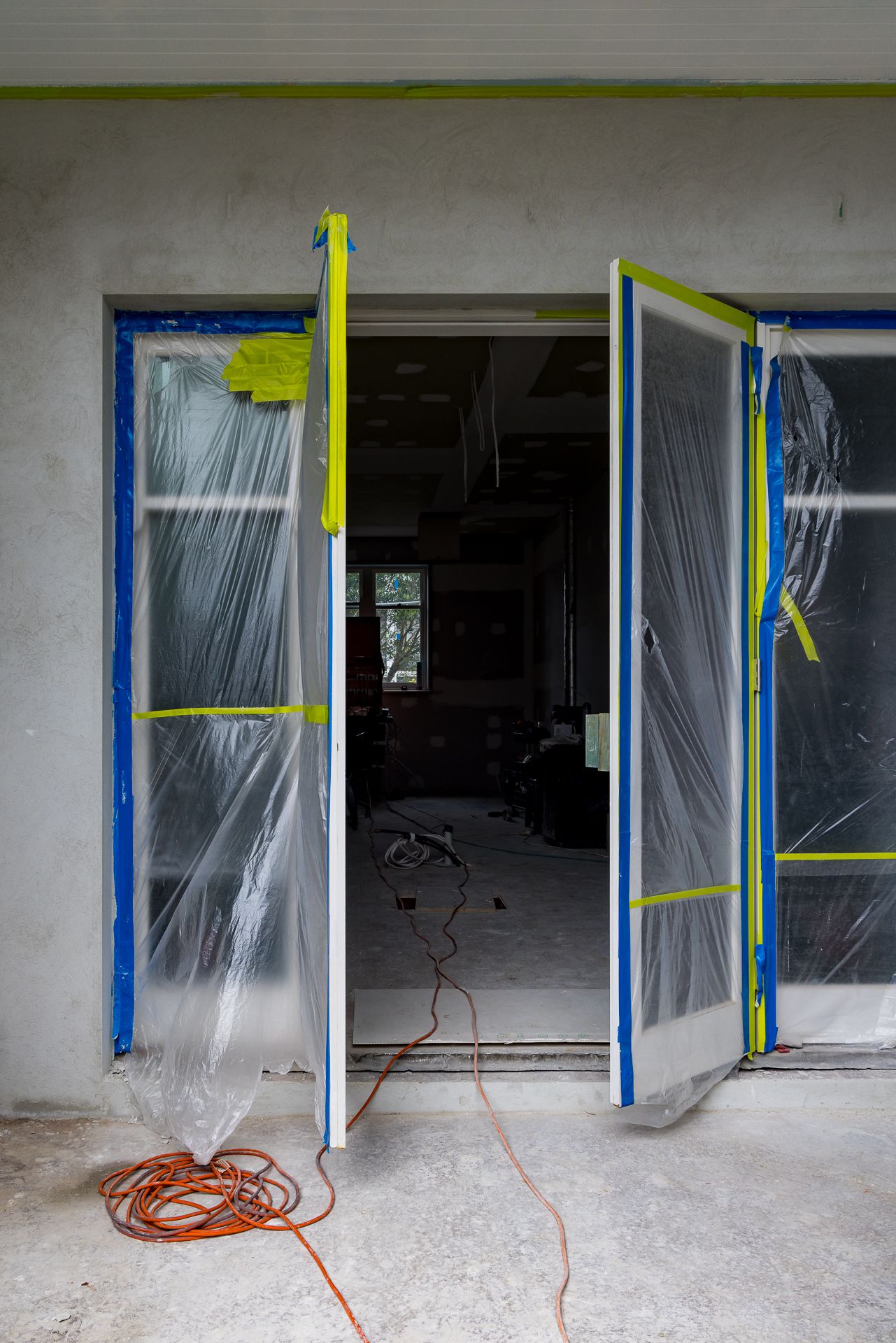 Open French doors wrapped in plastic sheeting and blue painter's tape at a home under construction, with an orange extension cord on the con