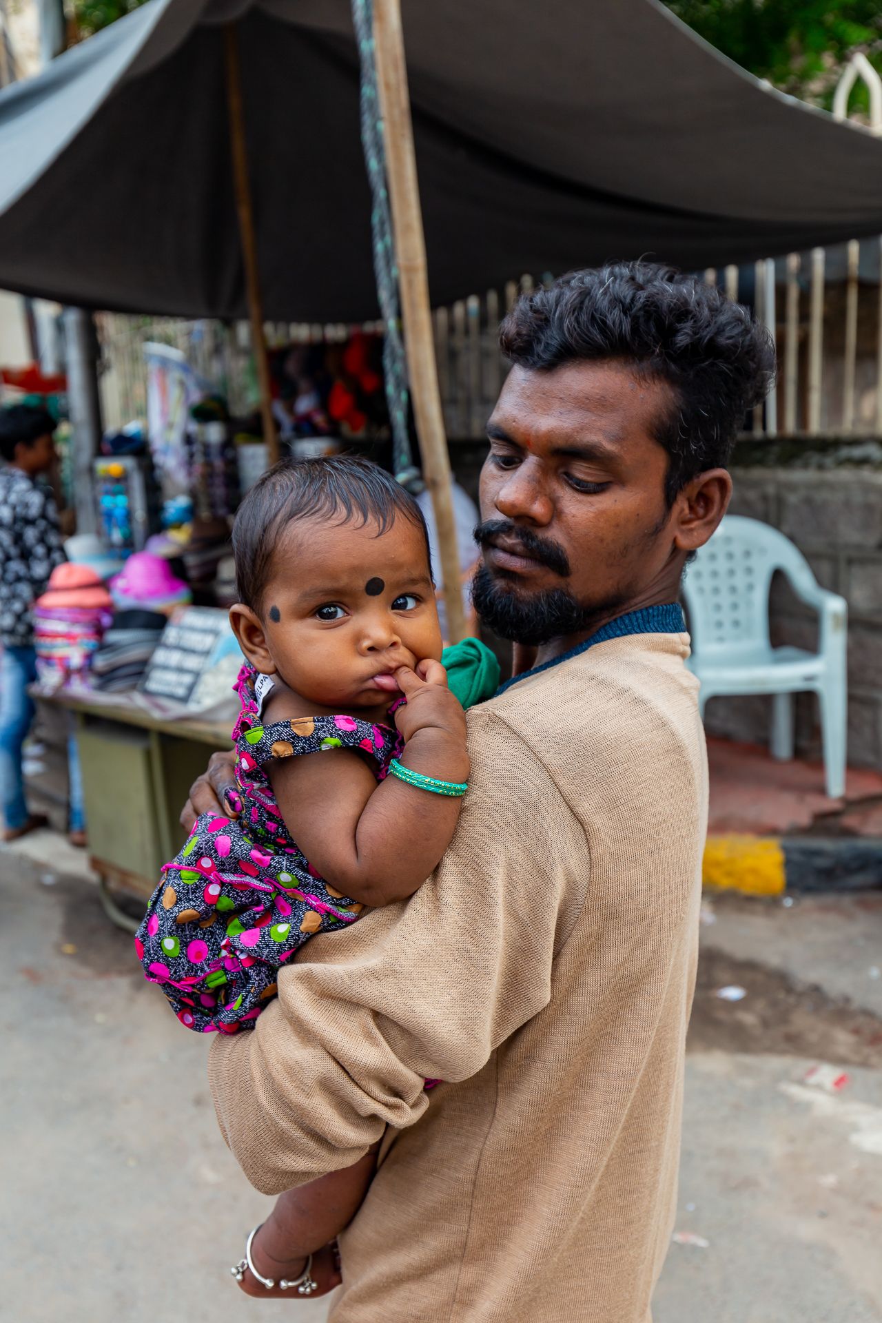 Man in a beige sweater holds a baby in a colorful floral dress at a busy street market with a tented stall behind them.