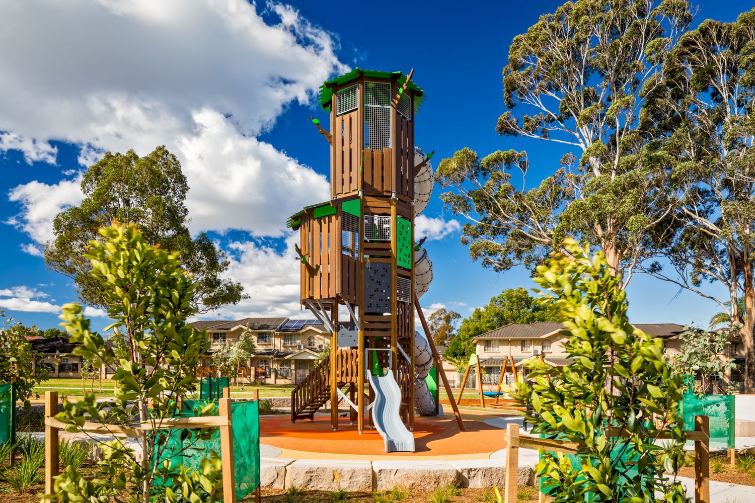 Modern wooden playground tower with a slide and climbing structure surrounded by trees and shrubs under a blue sky with clouds.