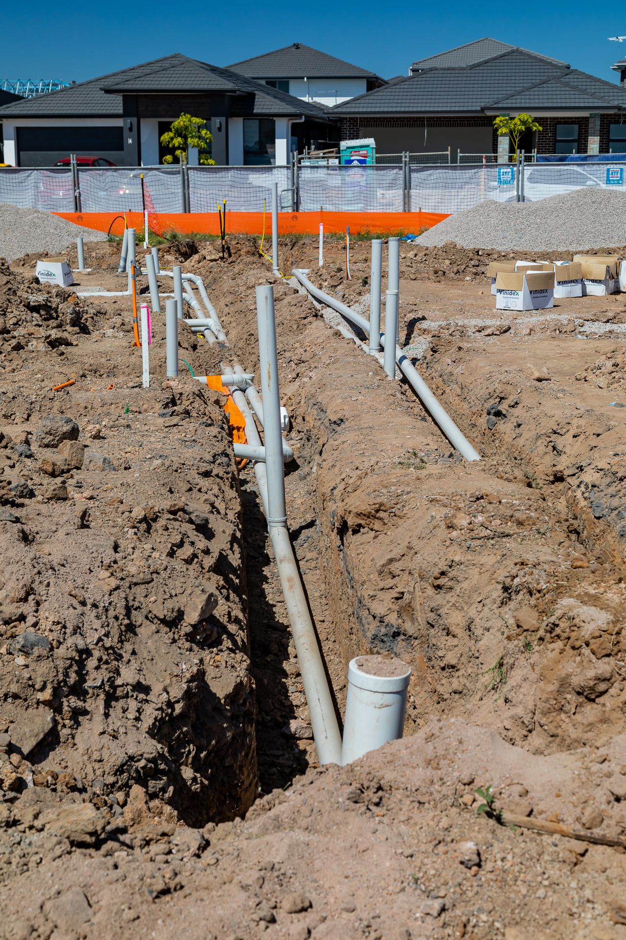 Open trench with grey drainage pipes and fittings installed in excavated soil in front of finished homes at a building site.
