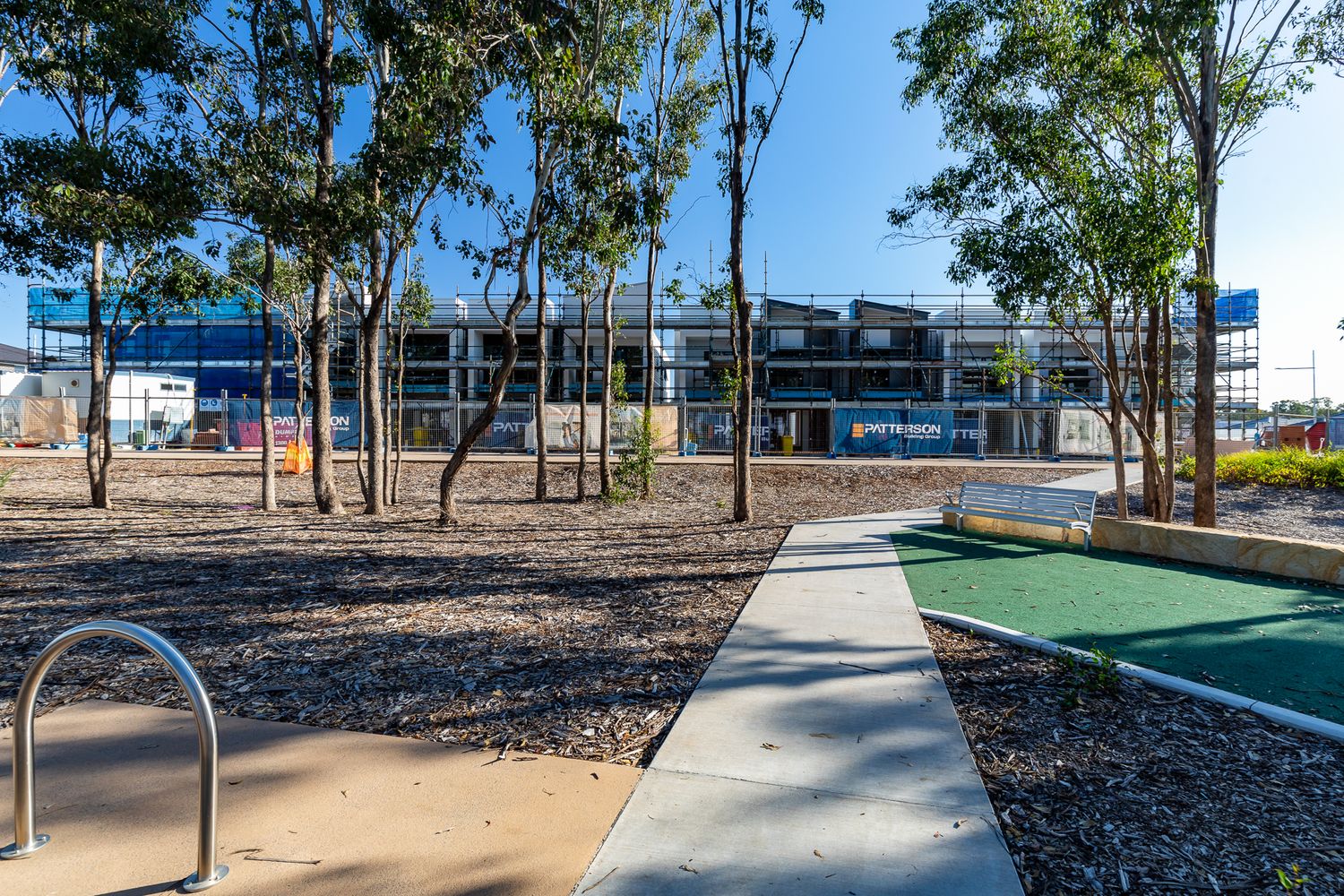 Landscaped park with trees, a concrete path and a bike rack, with a townhouse construction site visible in the background.