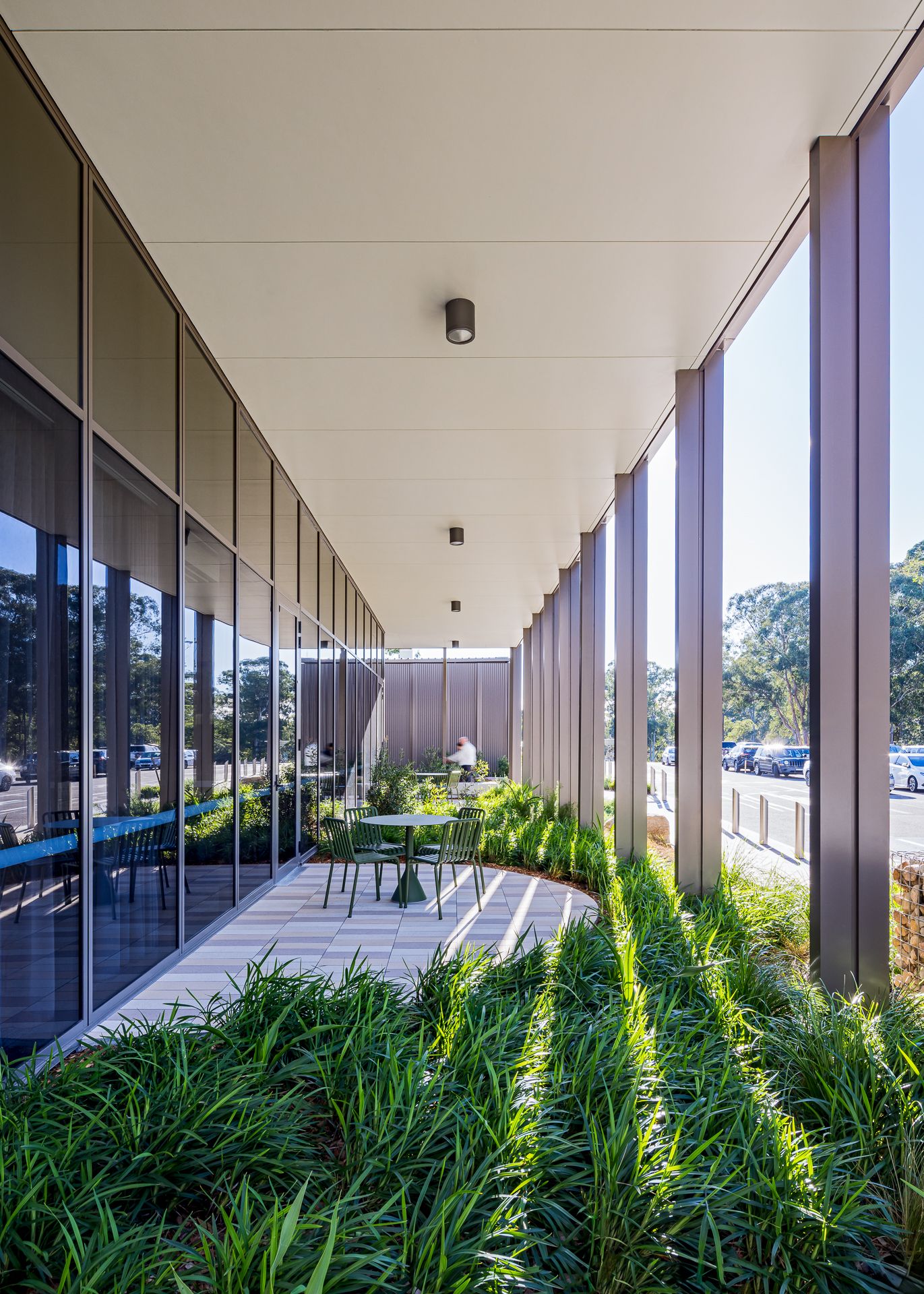 Covered outdoor walkway with a small bistro table and chairs beside floor-to-ceiling glass windows, lined with lush green grass-like plants.