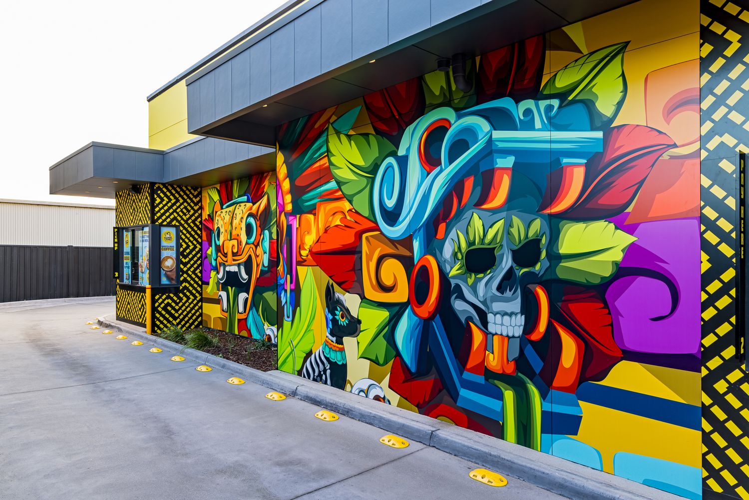 Colorful Aztec-inspired graffiti mural with a skull wearing a headdress and a jaguar face along a drive-thru restaurant wall.