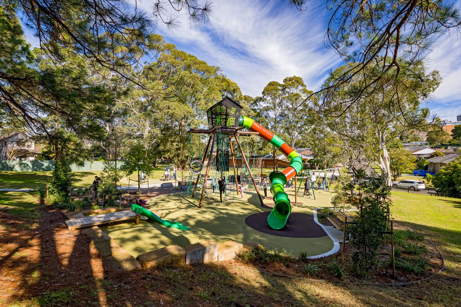Aerial view of a park playground with a treehouse-style tower, green and orange spiral slide, and surrounding shady gum trees.