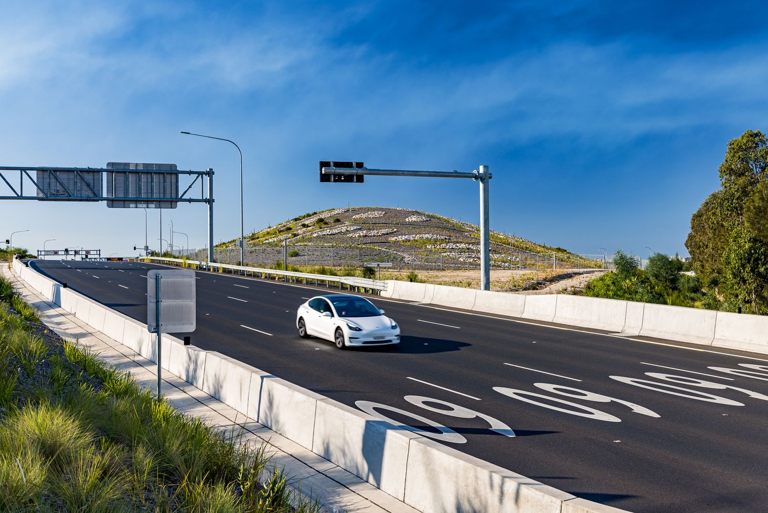 White electric car driving on a motorway toward a grass-covered landscaped hill and overhead traffic signage under a blue sky.