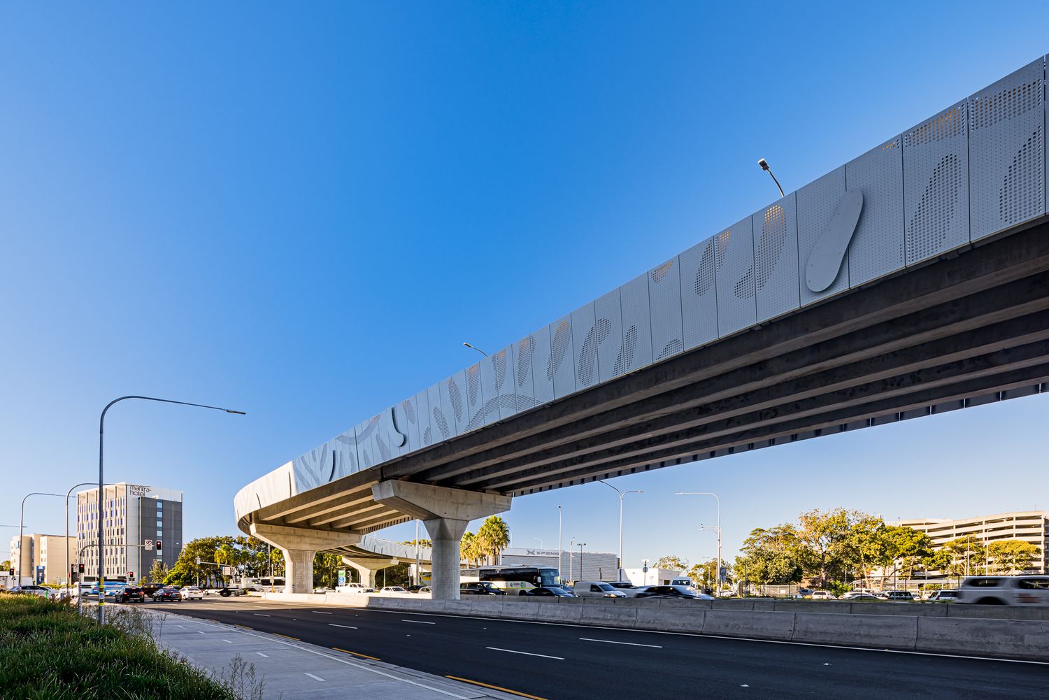 Long elevated transit viaduct with undulating panel cladding spans a wide road under a bright blue sky with trees and buildings nearby.
