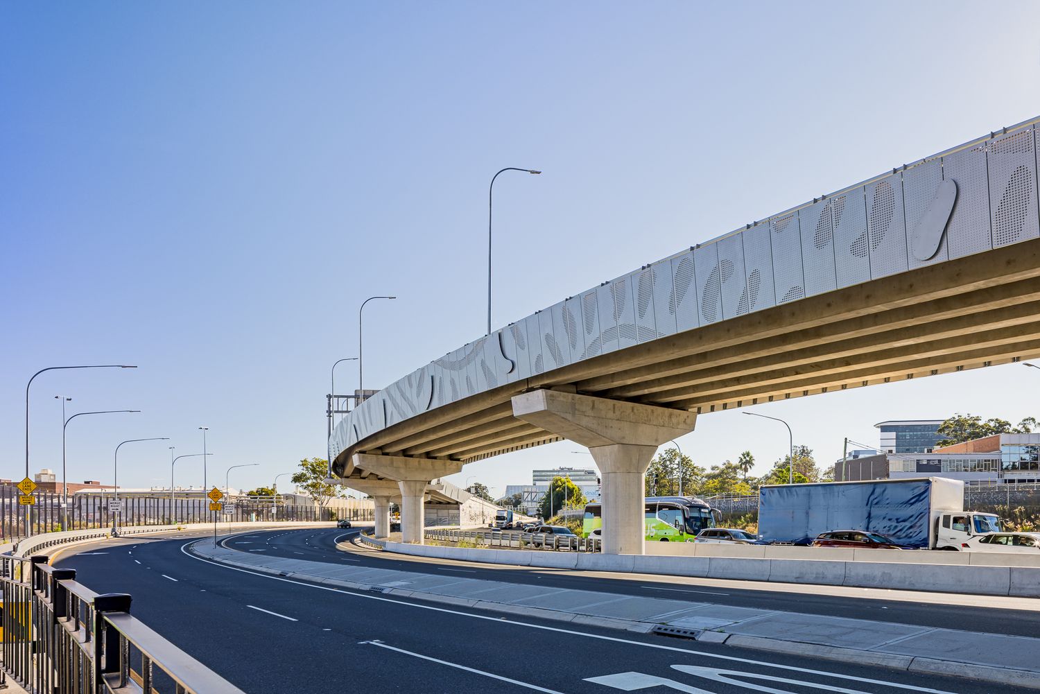 Curved elevated roadway on tall concrete pylons with decorative side panels above a road lined with streetlights and city buildings.
