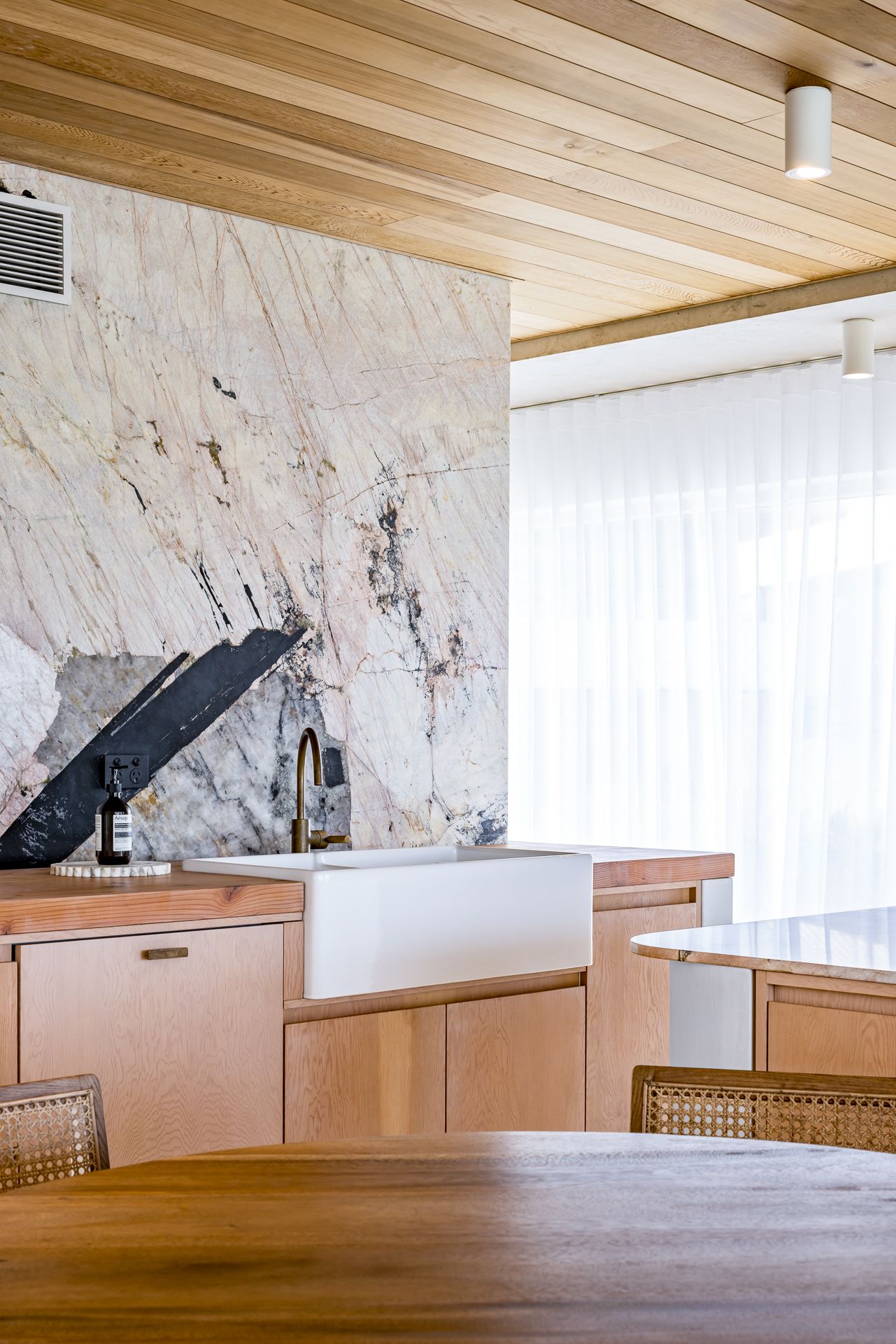 Kitchen detail showing a marble feature wall, white farmhouse sink with black tap, timber cabinets and a sheer-curtained window.