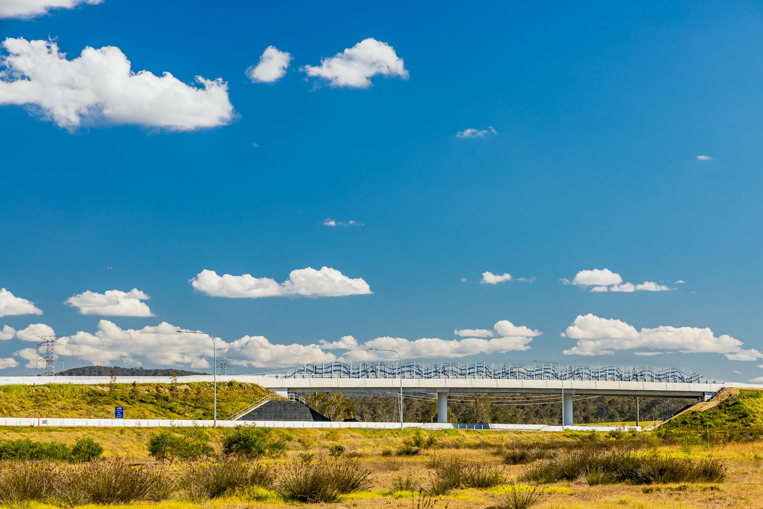 Distant motorway bridge spanning a dry grassy landscape beneath a blue sky with fluffy white clouds.