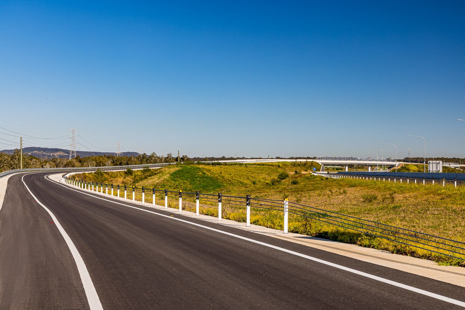 Newly paved curving road with white guide posts and safety fencing alongside green grassy hills under a bright blue sky.