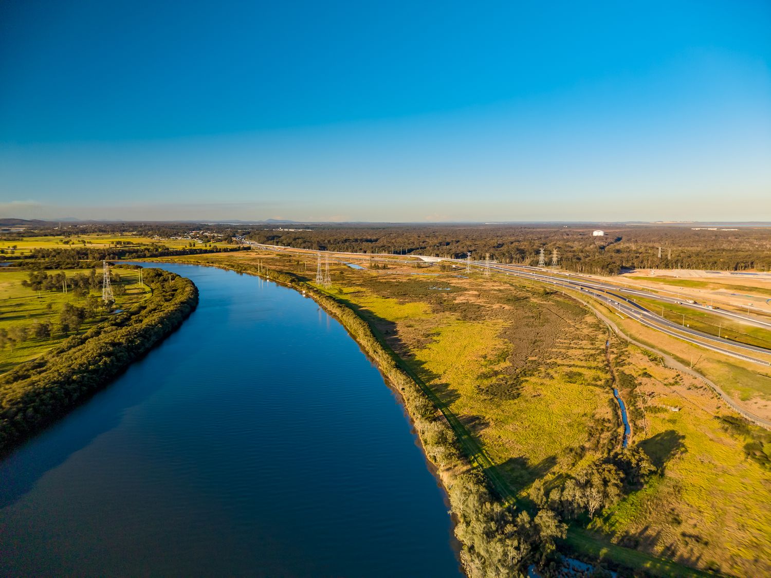 Golden-hour aerial view of a wide blue river curving beside open wetlands, power lines and a distant highway.