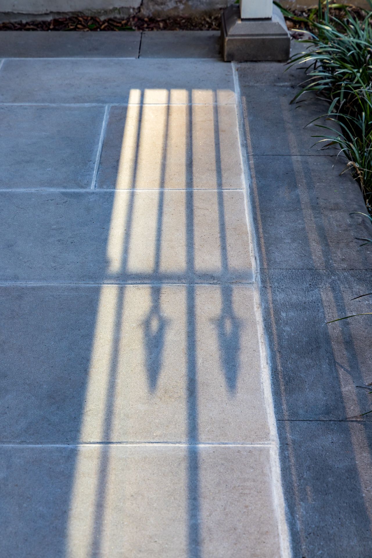 Sunlit shadow of a wrought-iron fence cast across a paved footpath beside a garden bed.