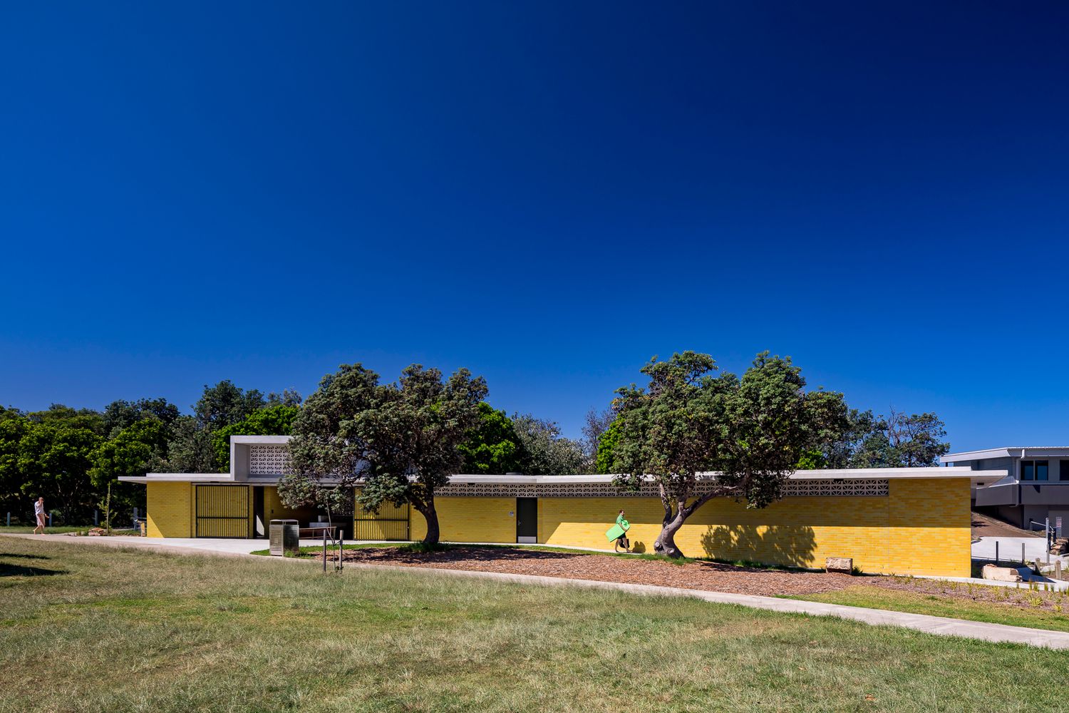 Wide view of a low yellow brick park pavilion with a flat roof, set among mature trees and open grass under a deep blue sky.