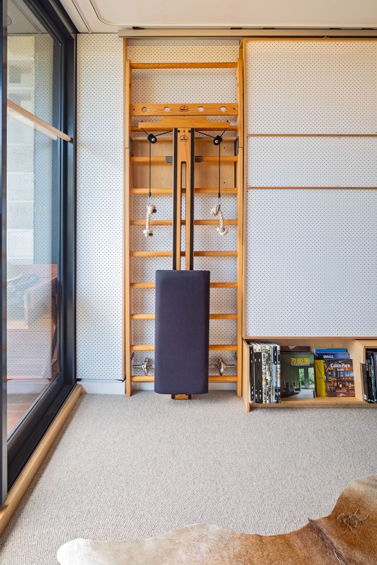 Wooden wall bars with a folded upright exercise bench in a bright home gym beside a glass door and a low bookshelf.