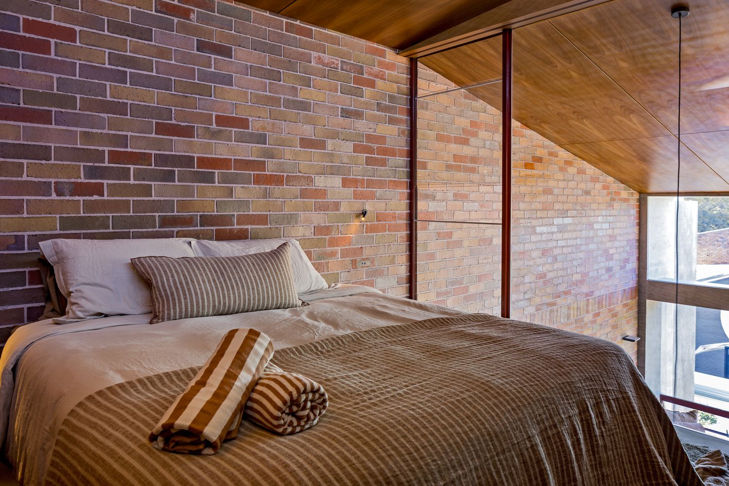 Attic-style bedroom with exposed multicolored brick wall, a timber ceiling, and a bed layered with striped bedding and rolled cushions.