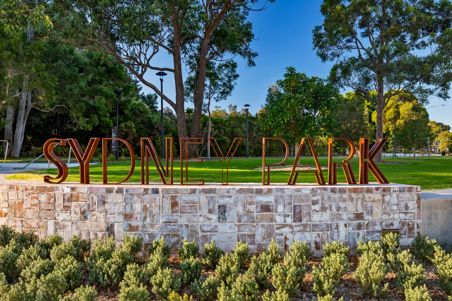 Large Sydney Park entrance sign in rust-coloured letters atop a sandstone wall with native shrubs and a green lawn behind.