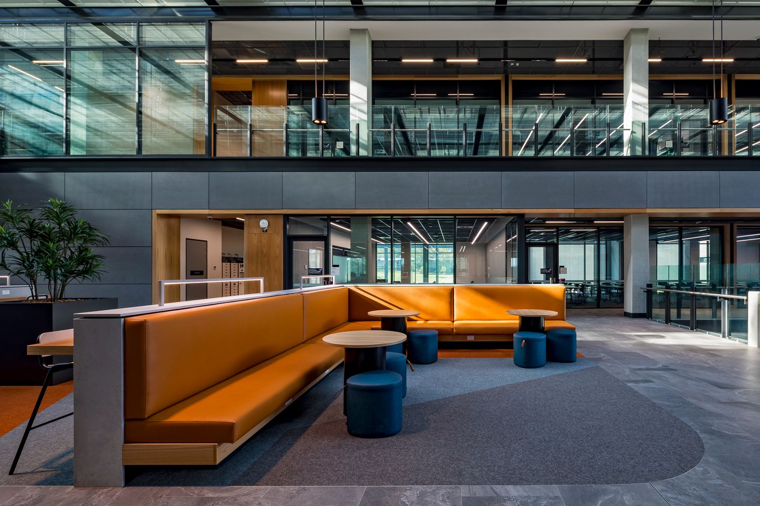 Contemporary TAFE interior lounge with orange built-in bench seating, round tables and blue stools beside floor-to-ceiling glass walls.