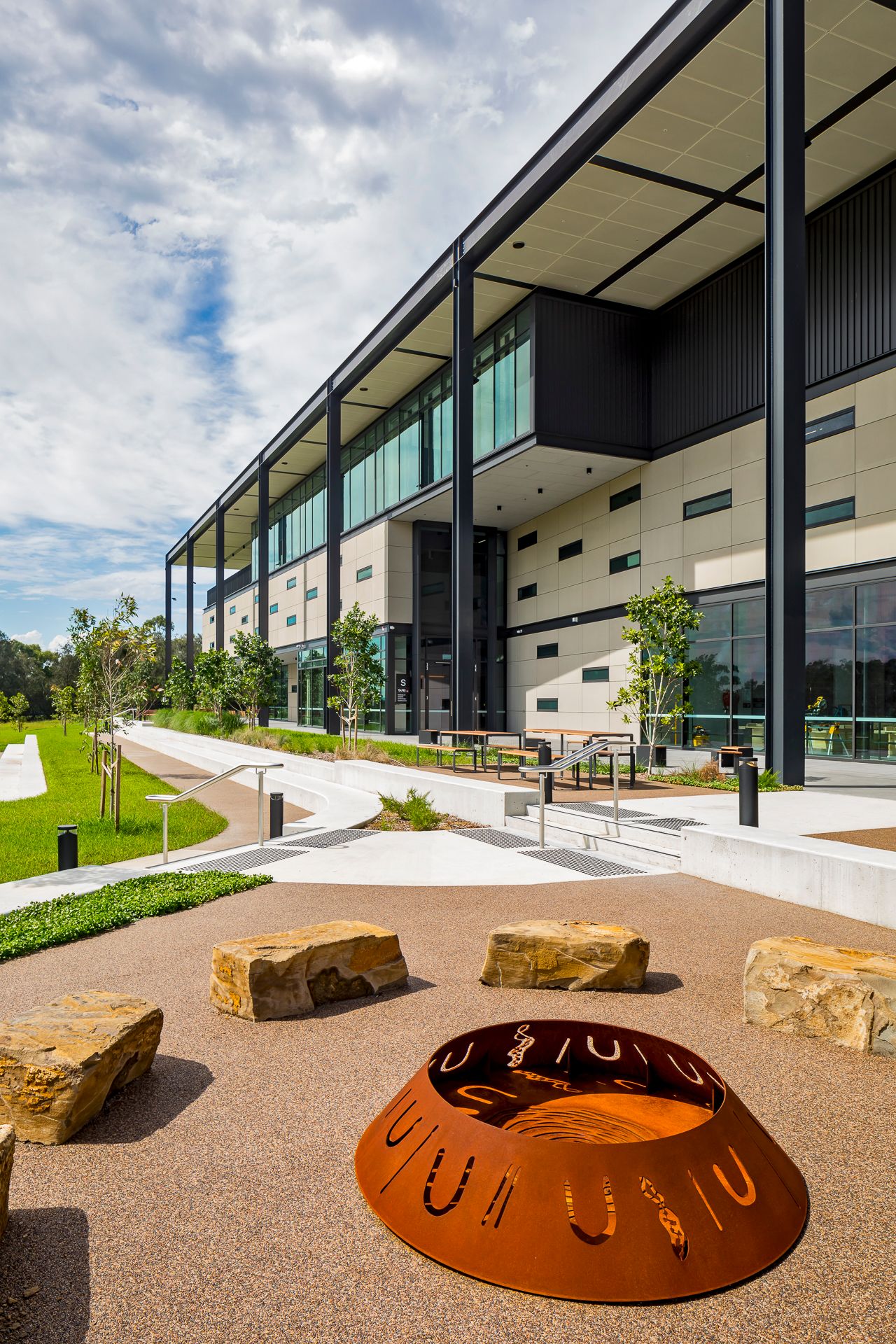 Multi-story modern campus building with glass facade and landscaped paths, with a laser-cut steel fire pit and boulders in the foreground.