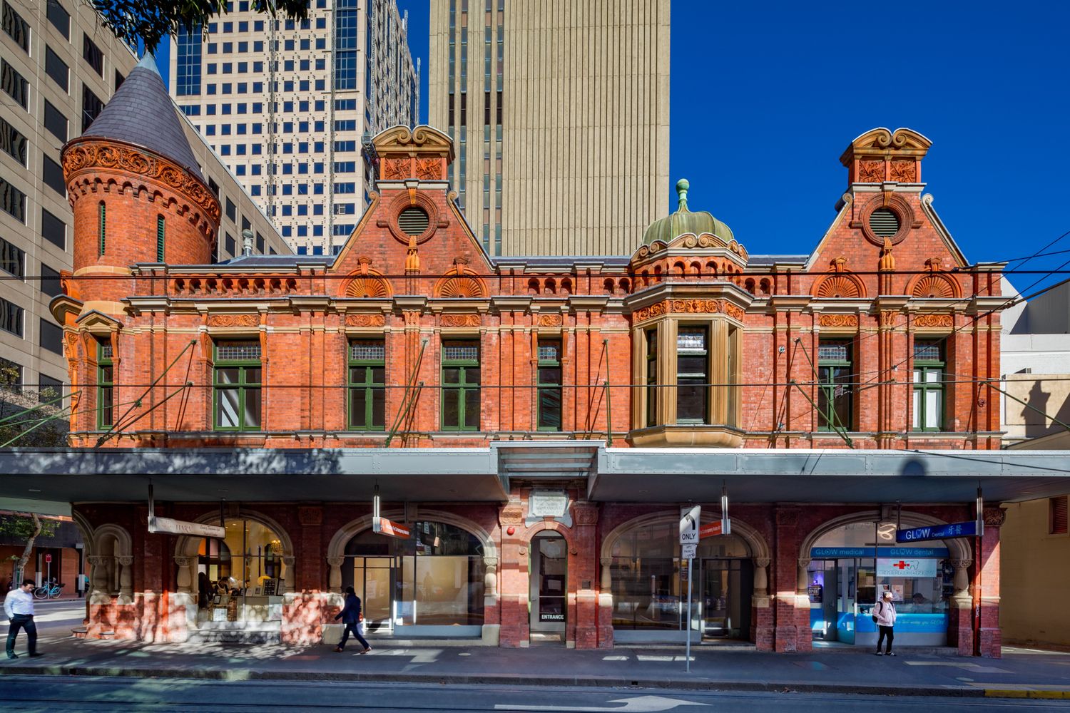 Victorian red brick corner building with decorative turrets and arched shopfronts, set against modern glass skyscrapers and blue sky.