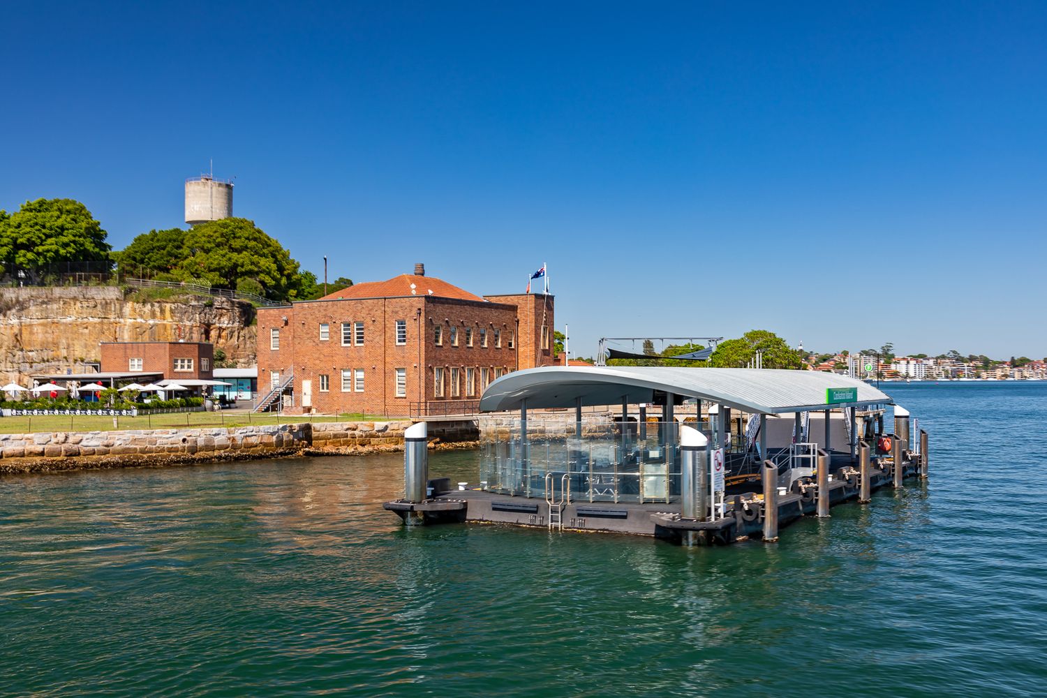Modern ferry wharf with a curved canopy beside a historic red-brick building and stone fortifications on a sunny Sydney harbour day.