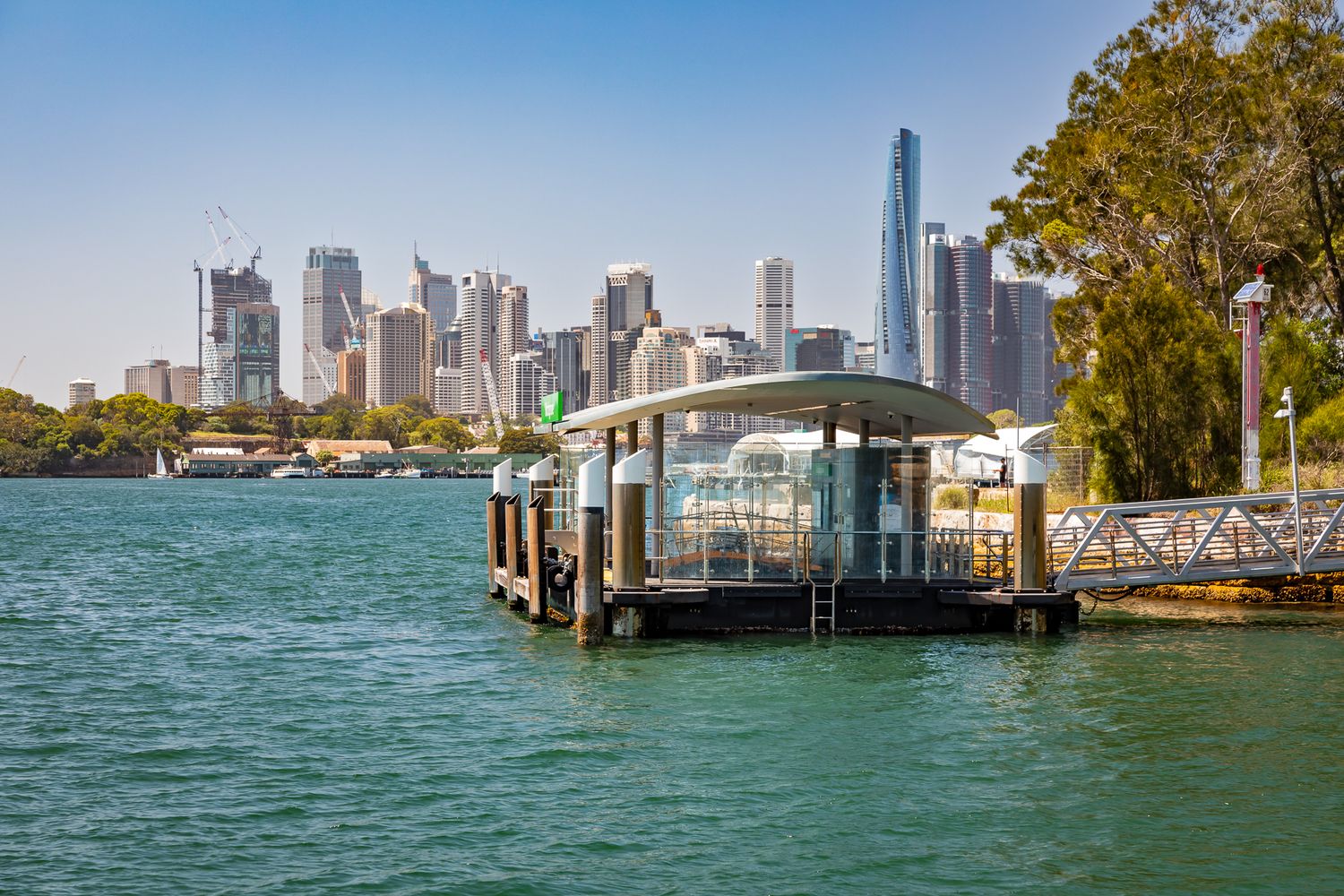 Ferry wharf with a curved canopy and gangway on turquoise harbour water, with the Sydney city skyline and tall towers in the background.