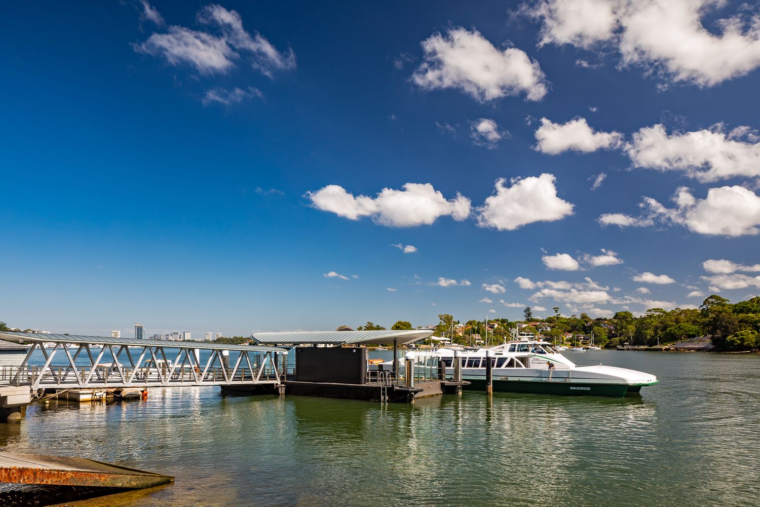 Modern ferry wharf with steel gangway and pontoon where a white ferry is moored on calm green harbour water under a bright blue sky.