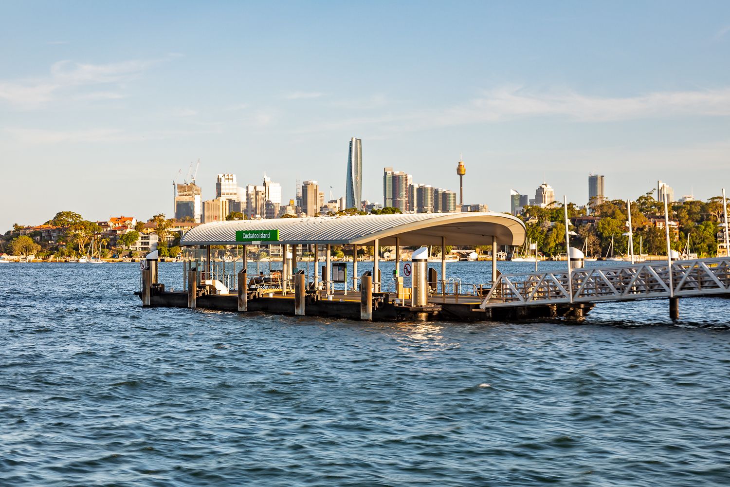 Covered ferry wharf on rippling harbour water, with city skyscrapers rising on the skyline behind it.