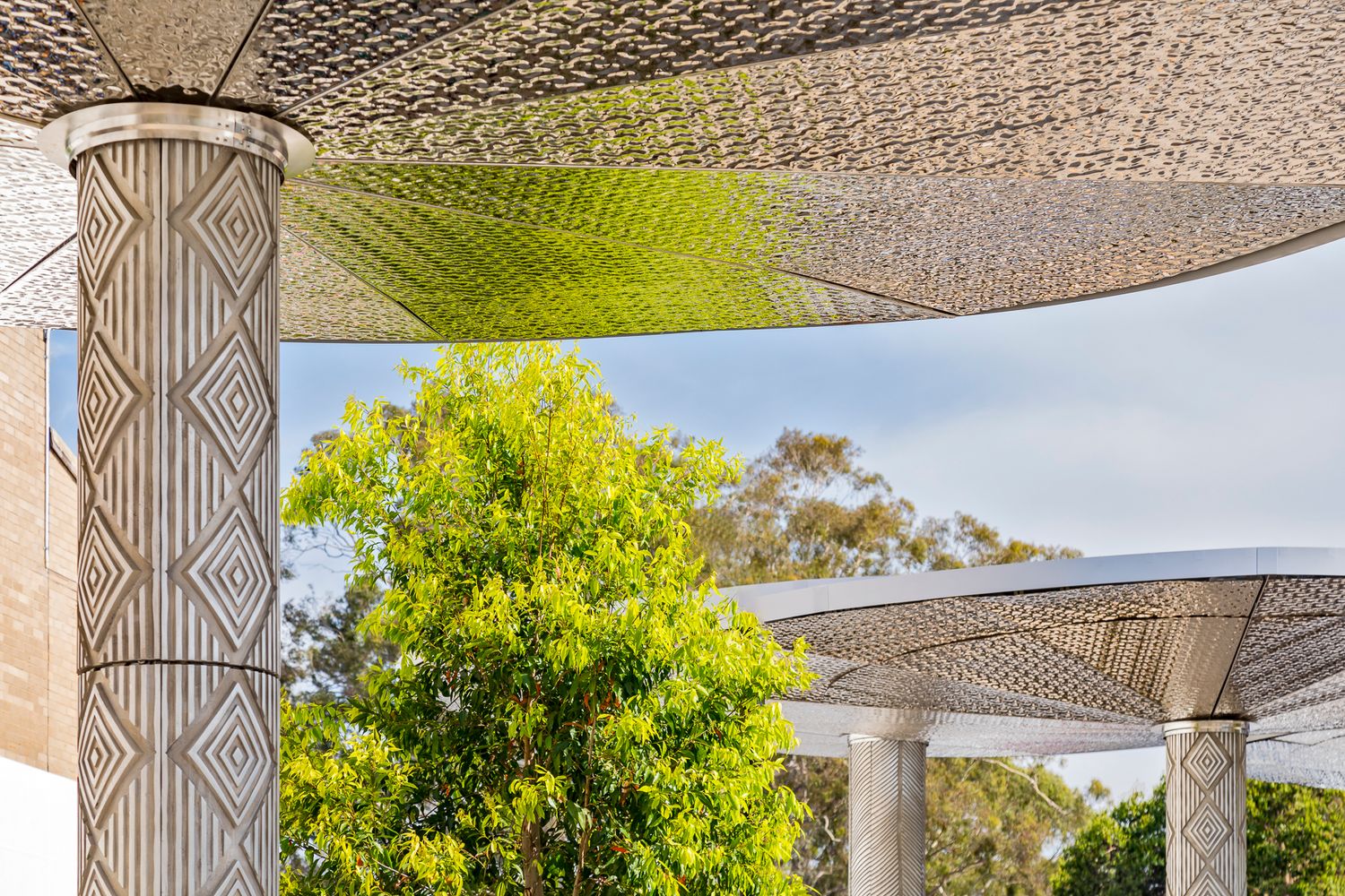 Close-up of an ornate diamond-patterned canopy column and lattice shade structure framing green trees against a bright blue sky.