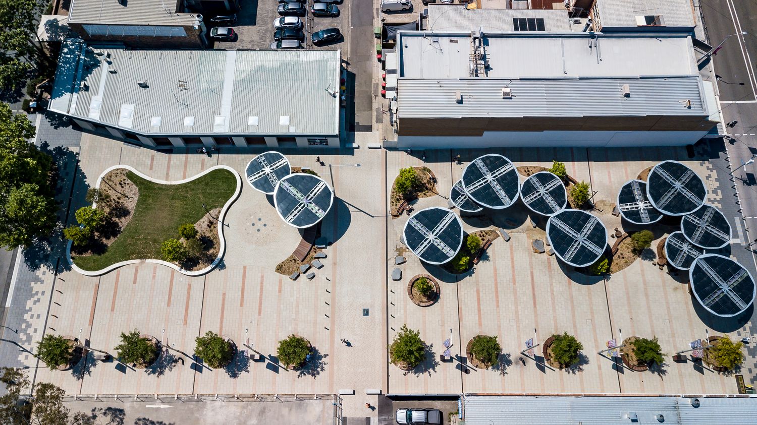 Bird's-eye view of a modern town square with solar canopies, a curved lawn and tree-lined paving between buildings.