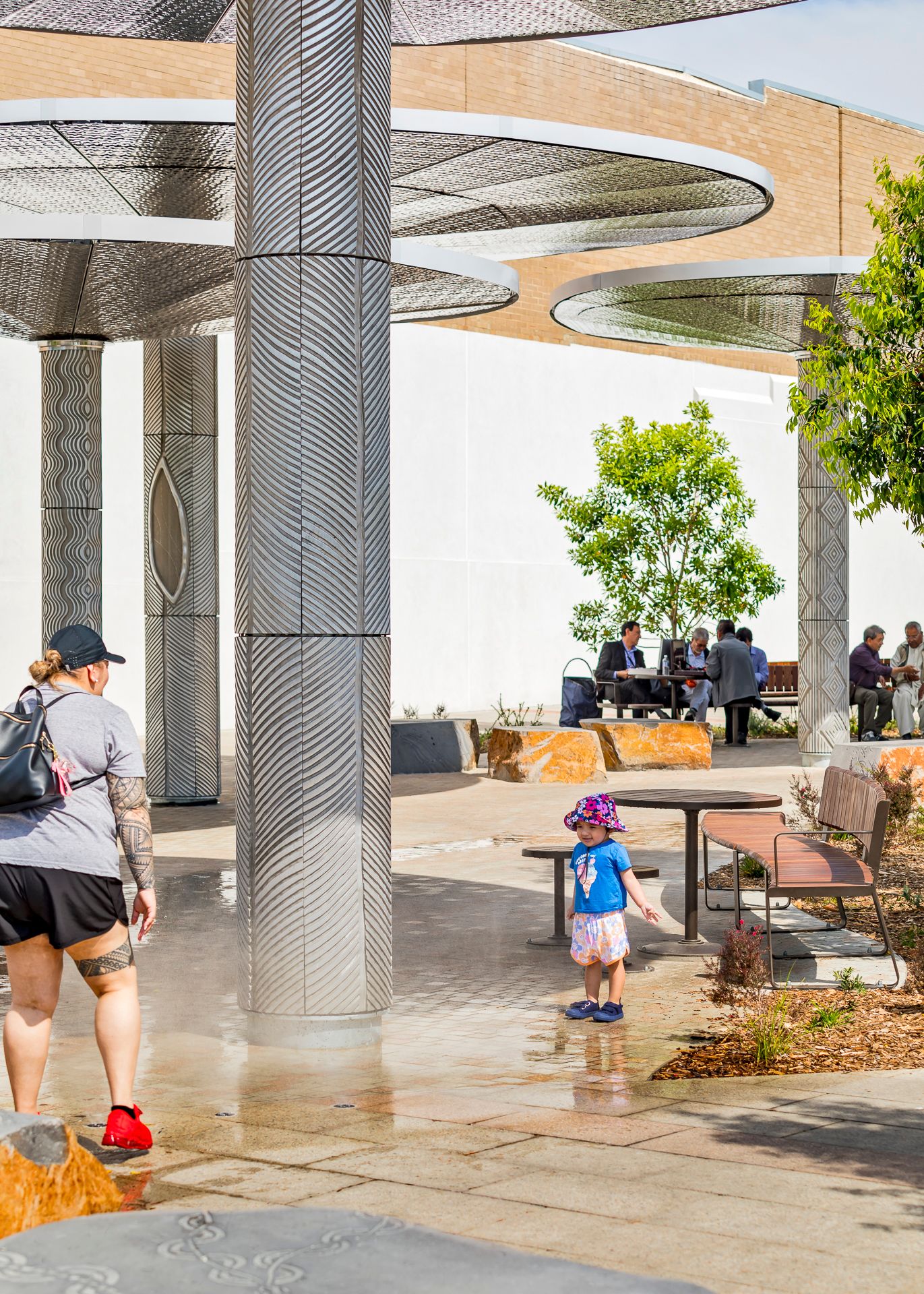 Mother and toddler in a pink hat walk through a plaza with patterned metal columns, benches and a small tree.