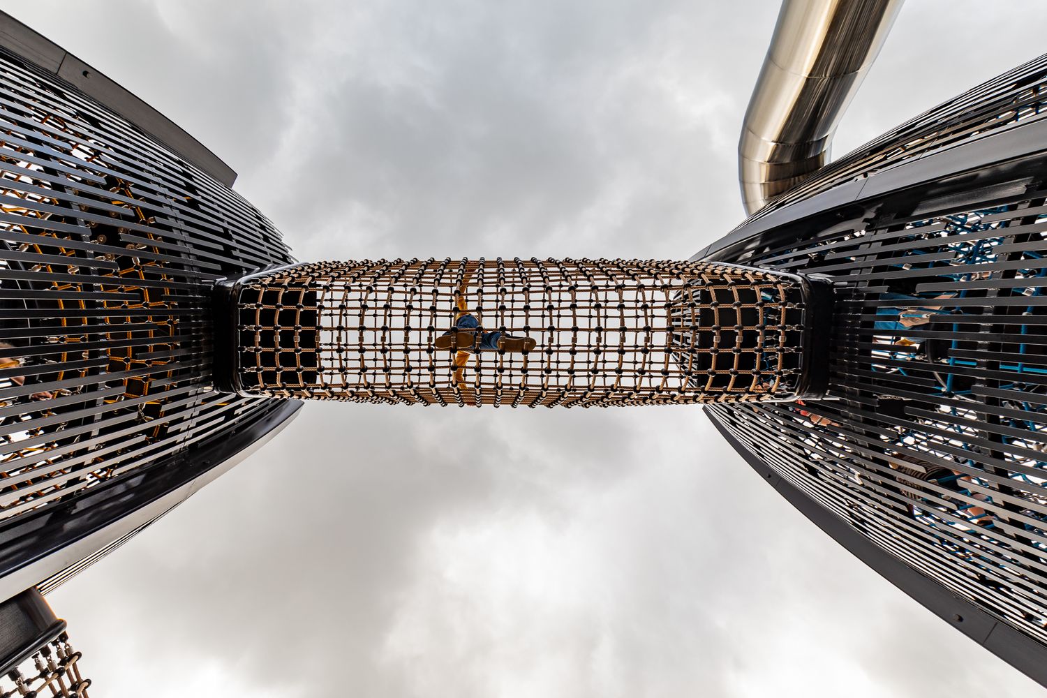 Low angle view of two dark steel climbing towers joined by a woven rope net tunnel against a cloudy sky.