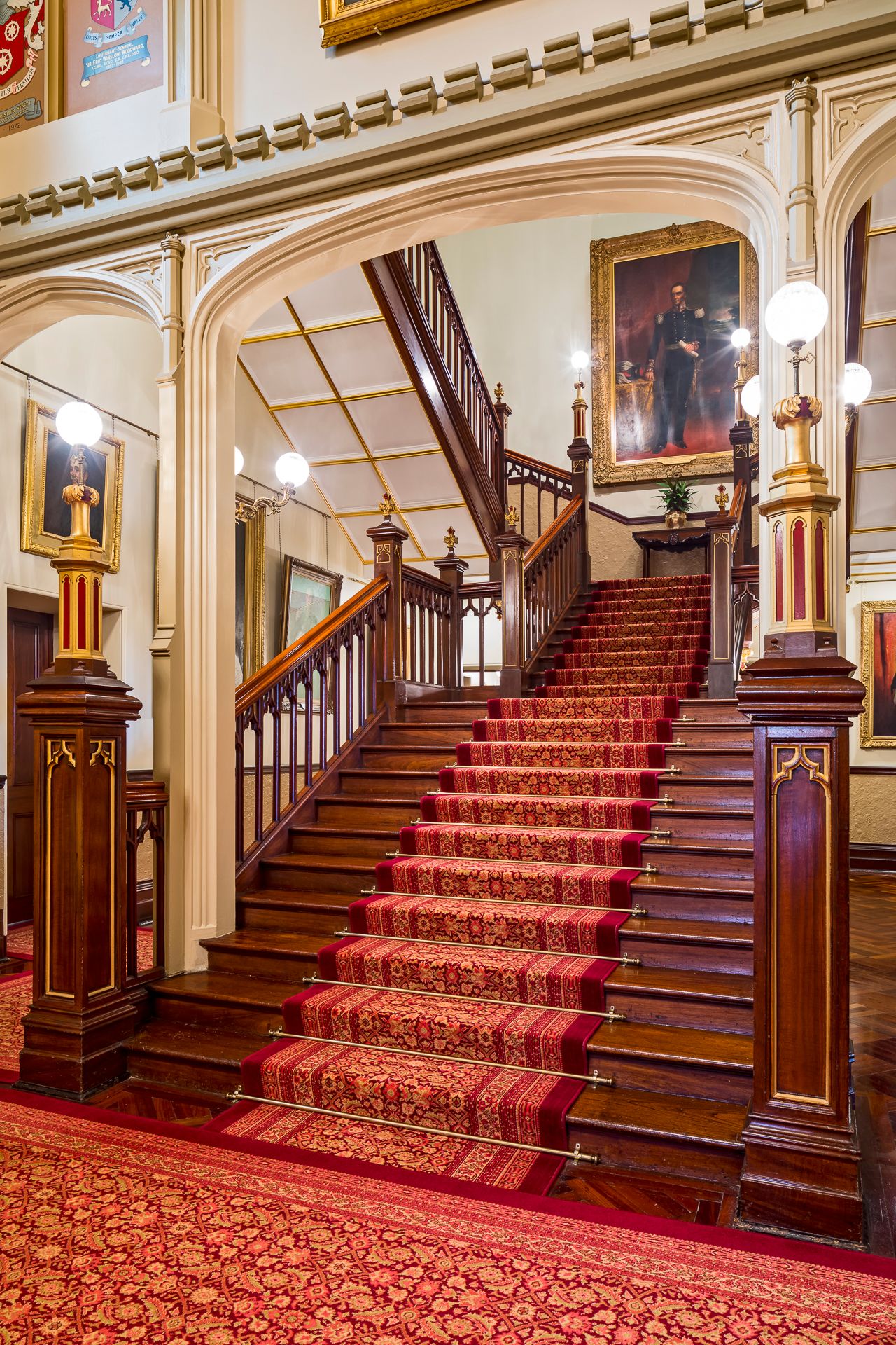 Elegant Victorian staircase with red carpet, ornate wooden balustrade, globe lamps and a framed portrait on the landing wall.