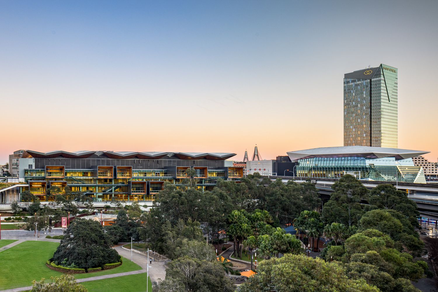 Dusk panorama of a city park with a timber-fronted convention centre and glass tower glowing under a pastel sunset sky.  ICC Sydney