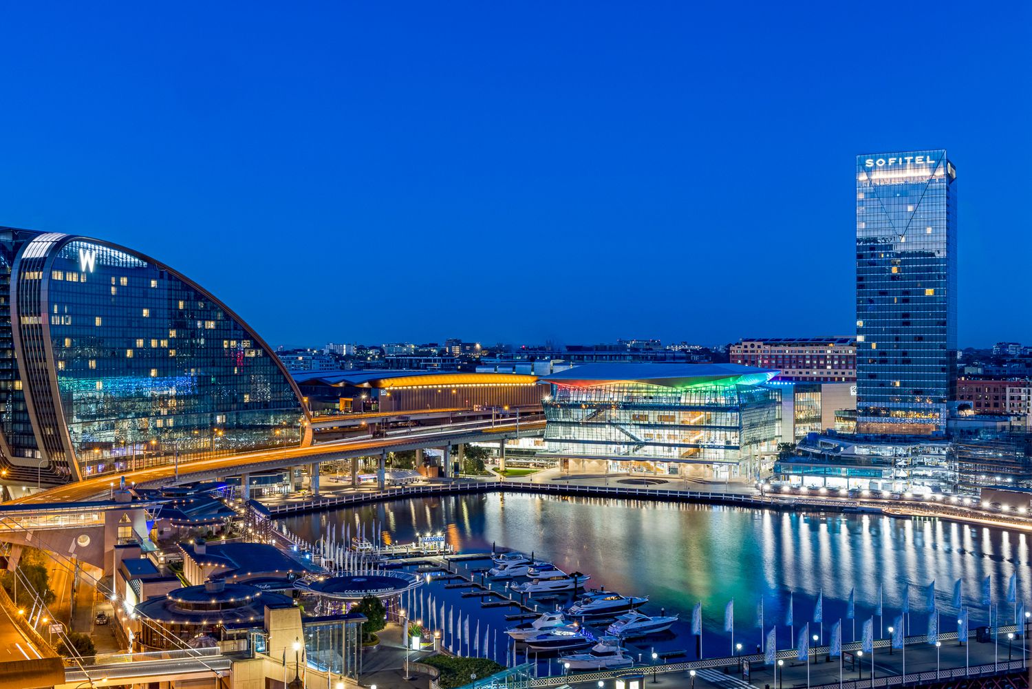 Illuminated waterfront skyline at dusk with a glass-domed building, a tall tower and moored boats reflecting on calm harbor waters.
