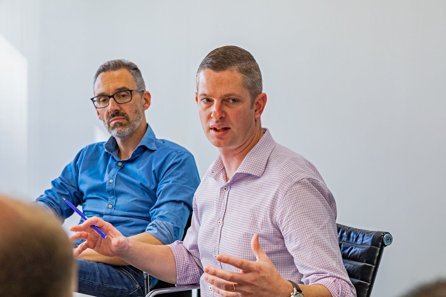 A man in a lilac checked shirt gestures while speaking at a meeting as a bearded colleague in glasses and a blue shirt listens beside him.