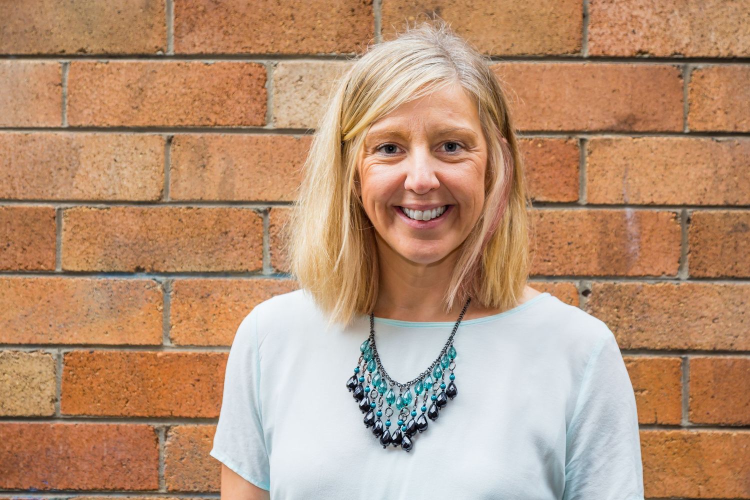 A smiling woman with blonde shoulder-length hair and a teal beaded necklace stands in front of a brick wall.