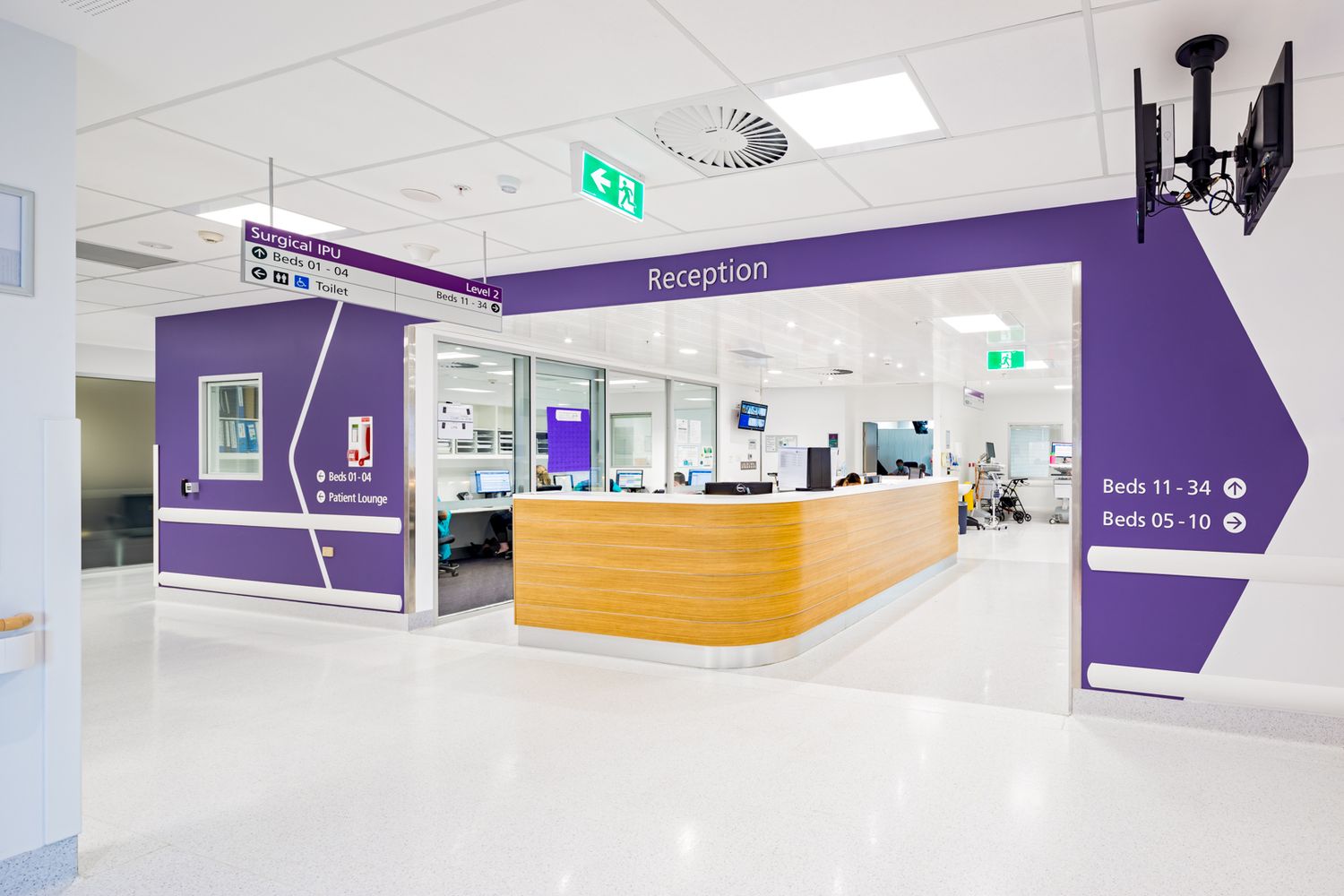 Hospital reception desk with timber counter and purple wayfinding signage showing bed numbers in a bright corridor.