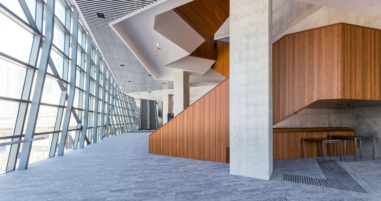 Interior concourse with floor-to-ceiling windows, concrete columns and a sculptural timber-panelled staircase.